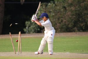 A cricket batsman gets bowled out as the red ball hits the stumps and dislodges the bails due to a batting mistake.